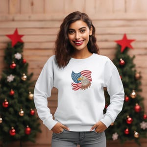 May include: White sweatshirt featuring an embroidered design of an American flag and a bald eagle. The eagle is brown and white, and the flag has red and white stripes with a blue field of stars. The sweatshirt is long-sleeved and has a crew neck.