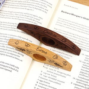 May include: Two wooden book page holders, one dark brown and one light brown, with butterfly and floral engravings. The dark brown holder has the name "Carmen" engraved. The light brown holder has the name "Eirene" engraved.