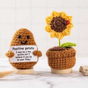 Puede incluir: Figura de patata tejida a ganchillo con un cartel que dice "Positive potato" y un girasol tejido a ganchillo en una maceta. El girasol tiene una mariquita. Ambos están sobre bases de madera.