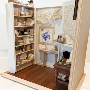 Może przedstawiać: Miniaturowa diorama gabinetu lub muzeum, z półką na książki, dziełami sztuki i małym stolikiem. Scena zawiera różne małe przedmioty, książki i elementy dekoracyjne, tworząc szczegółowe otoczenie. Podłoga jest z ciemnego drewna.