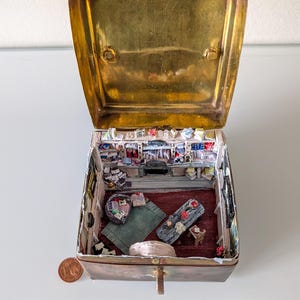 Puede incluir: Una pequeña caja de latón abierta que revela una detallada escena interior en miniatura. En el interior, una pequeña habitación está amueblada con objetos en miniatura, incluyendo una alfombra, un banco de madera y estanterías llenas de objetos en miniatura. Una moneda está al lado de la caja.