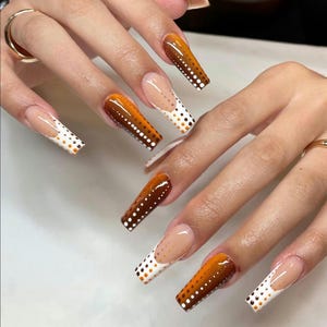 May include: Long, square-shaped artificial nails featuring a gradient design that transitions from a deep amber brown at the tips to a clear base near the cuticles. The nails are decorated with a pattern of small white and orange polka dots.