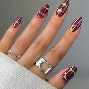 May include: Almond-shaped nails featuring a deep plum base colour with a light yellow starburst or grid pattern. Two silver-toned metal rings are worn on the fingers, one with a chain-link design and the other a wide band with a central cutout.