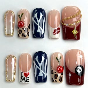 Puede incluir: Un conjunto de diez uñas artificiales con varios diseños. Los diseños incluyen una uña con borde dorado, un estampado de cereza y leopardo, una uña azul marino con un logotipo blanco, un diseño de labios y una uña con una cadena y una estrella. Las otras uñas presentan un diseño "I ❤️ ME", una cereza y un estampado de leopardo, y una bola 8.