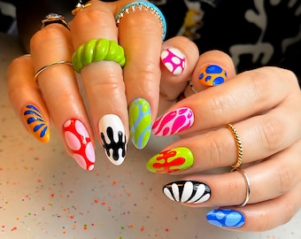 Könnte beinhalten: Nahaufnahme einer Hand mit langen, mandelförmigen Nägeln, die jeweils mit einem einzigartigen Design bemalt sind. Die Designs umfassen abstrakte Muster in leuchtenden Farben wie Pink, Orange, Grün und Blau. Die Hand hat auch mehrere Ringe.