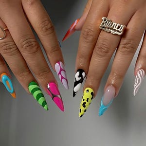 May include: Long stiletto-shaped artificial nails featuring a variety of vibrant designs including neon green stripes, hot pink with silver accents, black and white cow print, yellow leopard spots, blue swirls, and a silver ring reading Bianca.