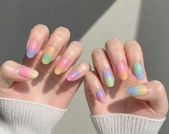 May include: Two hands displaying oval-shaped nails with a pastel rainbow gradient design. The nails transition from pink to yellow, green, and blue, creating a soft, colorful effect. The hands are set against a neutral background.