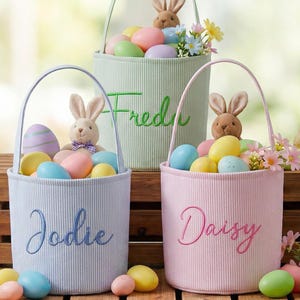 May include: Three pastel-colored Easter baskets with handles, each filled with colorful eggs, a plush bunny, and flowers. The baskets are labeled "Fred", "Jodie", and "Daisy". The baskets are on a wooden crate.