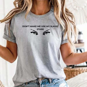 Könnte beinhalten: Graues T-Shirt mit dem Text "DON'T MAKE ME USE MY SLAVIC STARE" und einer Grafik von Augen. Das Shirt hat kurze Ärmel und einen Rundhalsausschnitt.