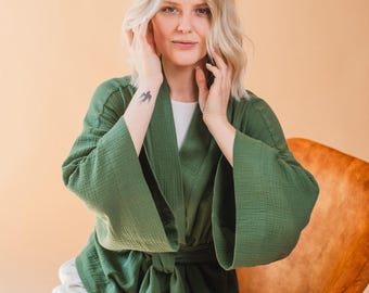 Op de afbeelding: Een groene kimono-achtige badjas met wijde mouwen en een bijpassende strikriem. De badjas is gemaakt van een textuurstof en wordt over een witte top gedragen. De persoon zit op een oranje stoel.