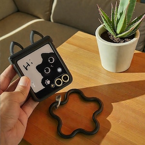 May include: Black phone case with cat ears and a cat graphic. The case has the word "Hi:" on it. A black star-shaped phone grip and a potted aloe vera plant are also visible on a wooden table.