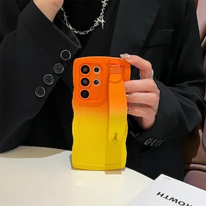 Könnte beinhalten: Eine orange-gelbe Handyhülle mit Farbverlauf und passender Handschlaufe. Die Hülle hat einen Kameraausschnitt mit drei Linsen und einen erhöhten Rand. Die Handschlaufe ist an der Seite befestigt.
