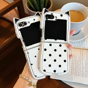 May include: Two white phone cases with black polka dots and cat ear designs. One case is open, revealing a black screen. The other case is closed, with black hearts. The cases are held by a person.