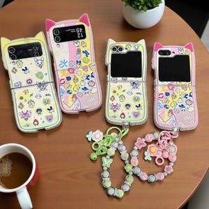 May include: Four brightly coloured phone cases with a cat ear design, decorated with cartoon characters and patterns. The cases are open and closed, displaying the phone screens. A beaded charm with flowers and butterflies is also present, with a cup of coffee.