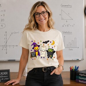 May include: A cream-coloured t-shirt featuring a graphic print of a cartoon pencil wearing a witch hat, a mummy, and a calculator with bat wings. The pencil holds a small chalkboard with the text "EQUATIONS ARE MY SPELL" written in white.