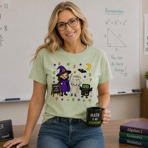 May include: A light green t-shirt featuring a graphic of a witch, a mummy, and a calculator with the text "NUMBERS ARE SPOOKY" surrounded by math symbols, a crescent moon, and a bat. The shirt is paired with a black mug that says "MATH IS MY SUPERPOWER."