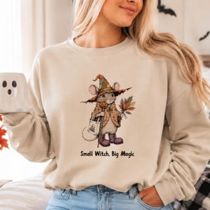 May include: A beige crewneck sweatshirt featuring a graphic illustration of a mouse dressed as a witch, wearing a patchwork hat, a cape, and purple boots. The mouse holds a small pumpkin staff and a bouquet of autumn leaves. Text reads: Small Witch, Big Magic.