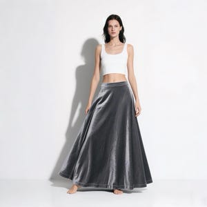 May include: A full-length, gray velvet skirt with a high waist and a flowing silhouette. The skirt is paired with a white crop top. The model is standing in front of a white background.
