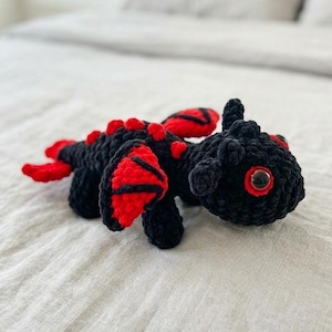Könnte beinhalten: Handgefertigter Häkeldraken in Schwarz und Rot. Der Drache hat rote Flügel, Stacheln und Augendetails. Das Plüschtier liegt auf einer weißen Oberfläche.