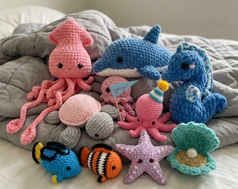 Puede incluir: Una colección de criaturas marinas de ganchillo que incluye un calamar rosa, un delfín azul, una tortuga marina, un pequeño pulpo rosa con una bandera azul que dice yay!, un caballito de mar azul, un pez azul, un pez payaso naranja, una estrella de mar morada y una almeja con una perla.