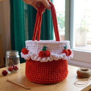 Könnte beinhalten: Handgefertigte Häkeltasche mit rotem Boden und weißem Oberteil, verziert mit Kirschmotiven. Die Tasche hat rote Henkel und einen gerüschten Rand. Ein Glas Kirschen und eine Häkelnadel sind zu sehen.