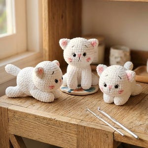 May include: Three handmade white crochet cat figurines with pink accents on a wooden surface. One cat is sitting upright, while the other two are lying down. Two silver crochet hooks are also visible.