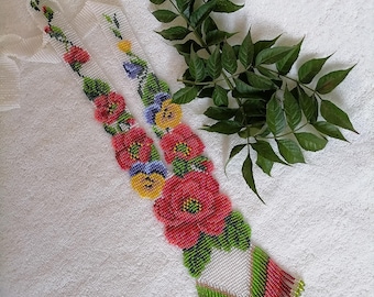May include: A beaded necklace featuring a floral design with red, yellow, and blue flowers. The necklace has a white base with green and red accents, and a fringed bottom. A green leafy branch is in the background.