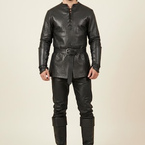 Könnte beinhalten: Ein schwarzes Leder-Ensemble, bestehend aus Tunika und Hose. Die langärmlige Tunika hat einen geschnürten Ausschnitt und einen Gürtel in der Taille. Die Hose ist eng anliegend, und das Outfit umfasst passende Lederstulpen und Stiefel.