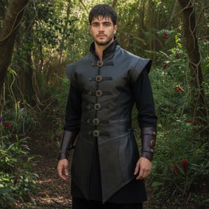 May include: A black leather vest with decorative buckles and shoulder pads. The vest is worn over a black long-sleeve shirt. The person is standing in a forest setting with green foliage and trees.