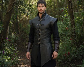 May include: A black leather vest with decorative buckles and shoulder pads. The vest is worn over a black long-sleeve shirt. The person is standing in a forest setting with green foliage and trees.