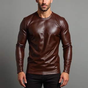 Può includere: Una camicia di pelle marrone a maniche lunghe con scollo tondo. La camicia ha un aspetto lucido e strutturato e viene indossata con pantaloni neri. L'immagine è su sfondo grigio.