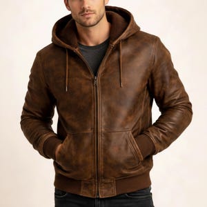 May include: A brown leather jacket with a hood and a zipper closure. The jacket has a distressed finish, a kangaroo pocket, and ribbed cuffs and hem. The interior lining is a darker brown color. The jacket is worn over a gray t-shirt.