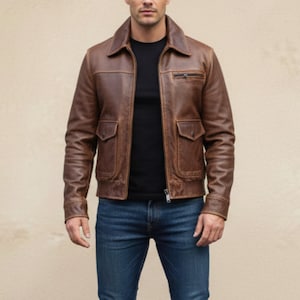 Könnte beinhalten: Braune Lederjacke mit Reißverschluss, zwei Vordertaschen und einer kleinen Brusttasche. Die Jacke hat einen klassischen Kragen und wird über einem schwarzen T-Shirt und blauen Jeans getragen. Die Jacke scheint eine mittlere Größe zu haben.