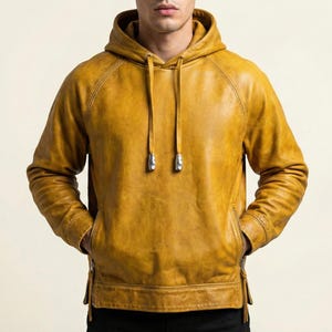 Könnte beinhalten: Senfgelber Leder-Hoodie mit Kapuze mit Kordelzug und Vordertaschen. Die Jacke hat einen Reißverschluss an jeder Seite der Taille und silberfarbene Metallakzente an den Kordeln.