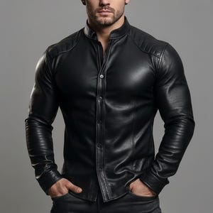 Può includere: Una camicia in pelle nera con colletto alla coreana e chiusura con bottoni. La camicia presenta dettagli trapuntati sulle spalle e maniche lunghe. Il modello indossa la camicia con le mani in tasca.