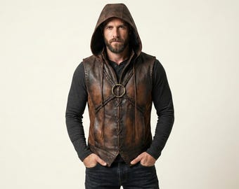 May include: A brown leather hooded vest with a bronze-colored ring closure. The vest has a distressed finish and is worn over a dark gray long-sleeved shirt and dark blue jeans. The hood is up, and the person is standing in front of a light background.