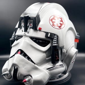 Puede incluir: Casco de piloto AT-AT blanco y plateado con detalles rojos y un símbolo imperial rojo. El casco presenta componentes mecánicos detallados, incluyendo una visera, tubos y una pieza de barbilla con punta roja. Artículo de colección.