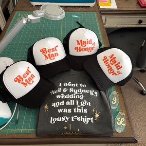 May include: A collection of black and white trucker hats with the words "Best Man" and "Maid of Honor" in orange script. A gray t-shirt with the text "I went to Neil & Sydney's wedding and all I got was this lousy t-shirt" is also present.