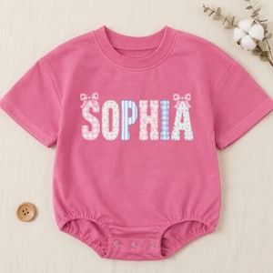 May include: A bright pink short-sleeve bodysuit featuring the name SOPHIA printed in large block letters across the chest. Each letter displays a unique pattern of pink gingham, blue stripes, or floral prints, accented by small pink bows on the ends.
