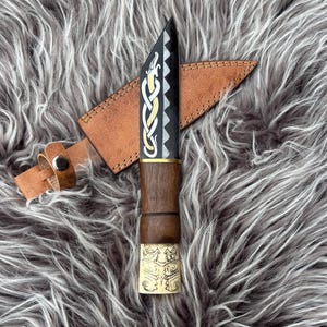 May include: A handcrafted knife with a black blade and a wooden handle. The blade features gold and silver decorative patterns. The knife is accompanied by a brown leather sheath with a belt loop.
