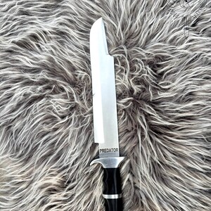 May include: A large, stainless steel knife with a black and silver handle. The word "PREDATOR" is etched on the blade. The knife is displayed on a gray, textured surface.