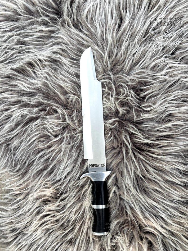 May include: A large, stainless steel knife with a black and silver handle. The word "PREDATOR" is etched on the blade. The knife is displayed on a gray, textured surface.
