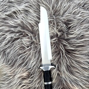 May include: A large, silver blade knife with a black handle and silver accents. The knife is set against a backdrop of gray and white faux fur. The knife has a wide, rectangular blade and a sturdy handle.
