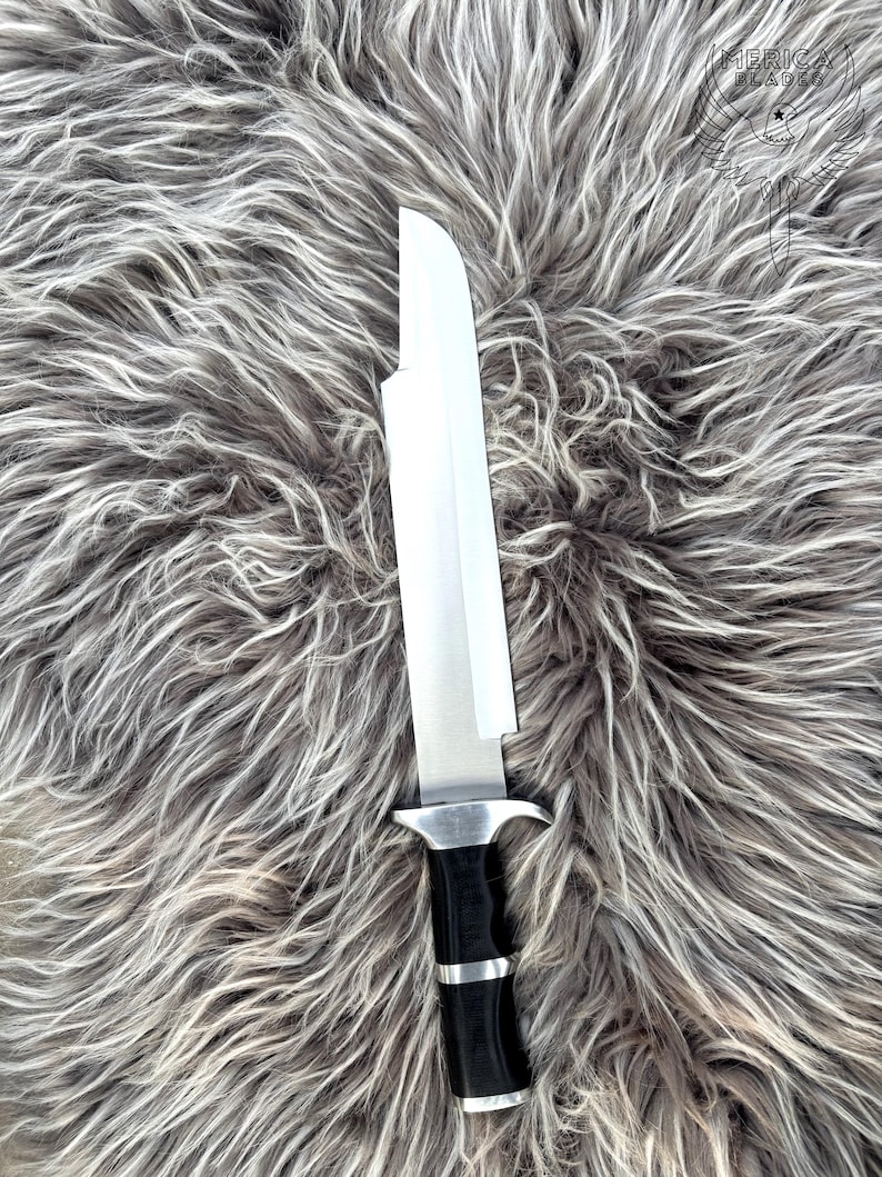May include: A large, silver blade knife with a black handle and silver accents. The knife is set against a backdrop of gray and white faux fur. The knife has a wide, rectangular blade and a sturdy handle.