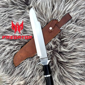 May include: A large hunting knife with a silver blade and a black handle, resting in a brown leather sheath. The word "PREDATOR" is printed on the blade. The knife is displayed on a textured, gray and white surface.
