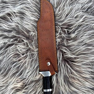 May include: A hunting knife with a brown leather sheath. The sheath is stitched with tan thread and has a silver snap closure. The knife handle is black with silver accents. The blade is partially visible.