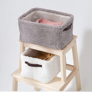 Puede incluir: Dos cestas de almacenamiento rectangulares, una gris y otra blanca, están sobre un taburete de madera. Las cestas tienen un exterior esponjoso y texturizado y asas marrones. La cesta gris contiene una prenda rosa, la blanca una prenda amarilla.