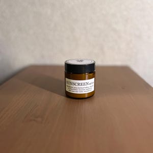 May include: A small, amber-colored jar of sunscreen with a black lid sits on a wooden surface. The label reads "SUNSCREEN spf 30" with a list of ingredients. The jar is centered in the frame, with a neutral background.