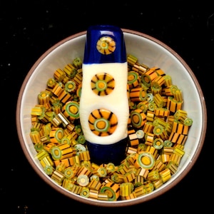 May include: A ceramic bowl filled with colorful glass beads and a decorative glass piece. The glass piece is white with orange and yellow circular designs and blue accents. The beads are yellow, orange, and green.