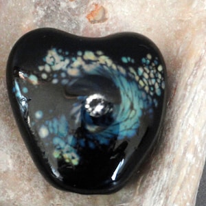 May include: A black heart-shaped glass bead with a swirling blue and white design. The bead has a shiny, glossy finish.